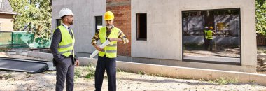 Güvenlik yelekleri içinde inşaat sürecini ve pankartı tartışan sadık mimar ve müteahhit.