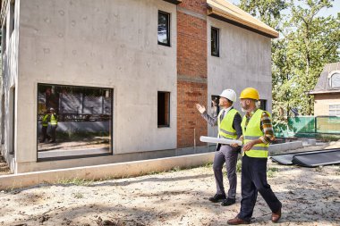 Güvenlik yeleklerinin mimarı ve müteahhidi elinde planlar tutuyor ve inşaat sürecini tartışıyor.