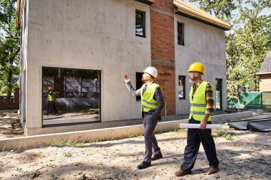 Güvenlik yeleklerinin mimarı ve müteahhidi elinde planlar tutuyor ve inşaat sürecini tartışıyor.
