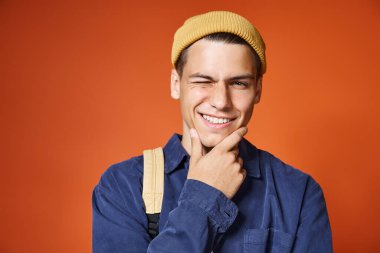 Sarı şapkalı yakışıklı bir gencin portresi. Gri gözlü. Göz kırpıyor ve elleriyle çenesine dokunuyor.