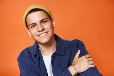 portrait of handsome young man in casual outfit and yellow hat smiling against terracotta background clipart