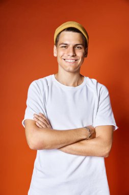 Beyaz tişörtlü, sarı şapkalı ve kollarını kavuşturmuş gülümseyen çekici bir genç adam portresi.
