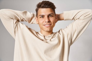 charismatic man in white hoodie smiling and putting hands behind head against light background clipart