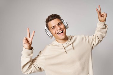 cheerful student in headphones smiling and showing sign peace with hands against light background clipart