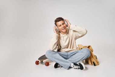 attractive man in headphones sitting with backpack and skateboard and listening to music clipart