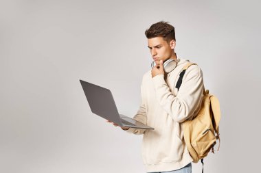 Sırt çantalı kulaklıklı düşünceli öğrenci gri arka planda dizüstü bilgisayar kullanıyor.