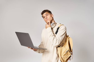 thoughtful man in headphones with backpack networking to laptop and looking up on grey background clipart