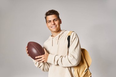 handsome student in his 20s with backpack holding rugby ball on grey background clipart