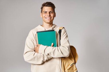 smiling student in 20s with backpack holding notes with hands against grey background clipart