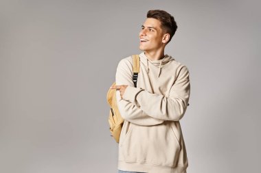Sırt çantası ve katlanmış kolları olan çekici bir genç adam gri arka planda parmaklarını gösteriyor.