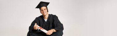 Mezuniyet elbisesi ve şapkalı mutlu adam pankartı. Oturmuş ve diplomasını elleriyle tutuyor.