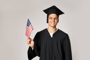 Mezuniyet kıyafetli yakışıklı genç adam, gri arka planda Amerikan bayrağıyla poz veriyor.