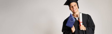 Diploma ve Avrupa bayrağıyla gri arka planda poz veren mezuniyet üniformalı bir adam.