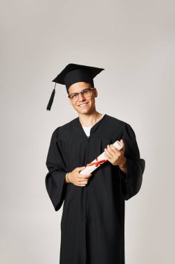 confident student in graduate outfit with vision glasses smiling with diploma in hands clipart