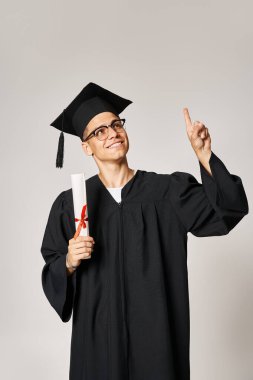 charming student in graduate outfit with vision glasses pointing finger to up with diploma in hand clipart