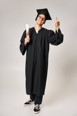 Mezuniyet kıyafetli, vizyon gözlüklü, elinde diplomayla elini havaya kaldıran neşeli genç bir adam.