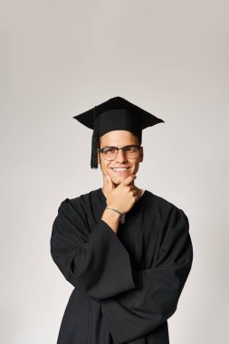 smiling student in graduate outfit and vision glasses touching hand to jawline on grey background clipart