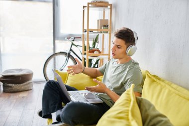 Evde sarı koltuklu kulaklıklı adamın profili laptopta online toplantıyla konuşuyor.