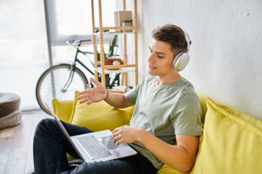 Evde sarı koltukta kulaklıklı çekici bir genç dizüstü bilgisayarla online toplantıyla konuşuyor.