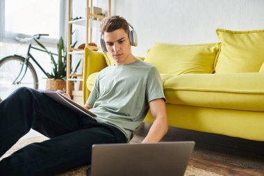 Kulaklıklı genç adam sarı kanepenin yanında dizüstü bilgisayarda çalışıyor ve not yazıyor.