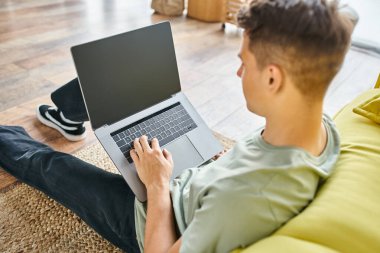 Evdeki sarı kanepenin yanındaki yerde oturan yakışıklı genç adam dizüstü bilgisayarla arkadan iş çeviriyor.