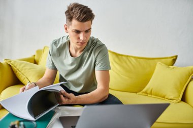 young man in yellow couch concentration doing coursework with notes and laptop on coffee table clipart