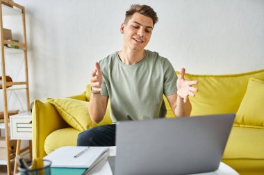 Sarı kanepede dizüstü bilgisayarlı çekici bir öğrenci ve sehpadaki notlar online toplantıyla konuşuyor.