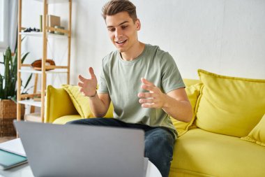 Sarı kanepede laptopuyla sehpanın üzerinde neşeli bir öğrenci çevrimiçi toplantıyla konuşuyor.