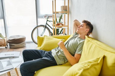 sideways of weary man in his 20s putting hand behind head and leaning on yellow couch with glasses clipart