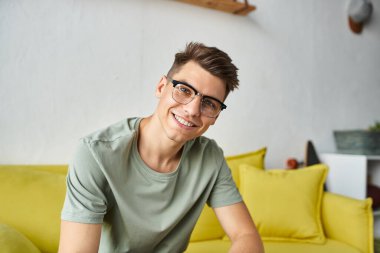 cheerful student in his 20s with vision glasses smiling on yellow couch in living room clipart