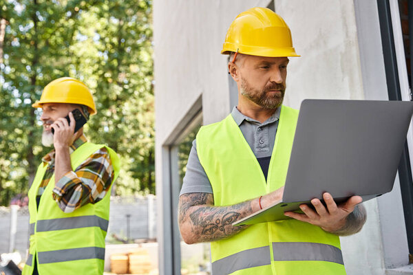 привлекательный строитель с татуировками, работающий на ноутбуке, пока его коллега разговаривает по телефону во время работы