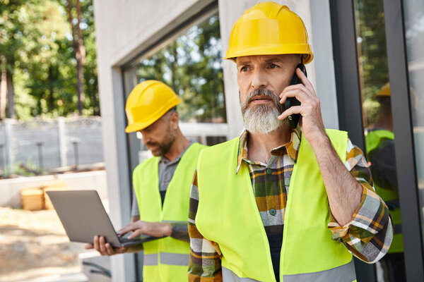 привлекательный строитель с татуировками, работающий на ноутбуке, пока его коллега разговаривает по телефону во время работы