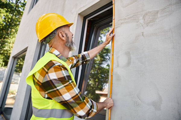 good looking hardworking construction worker with beard measuring window with tape, cottage builder
