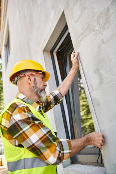 stock image handsome cottage builder in safety vest and helmet measuring window with tape while on site