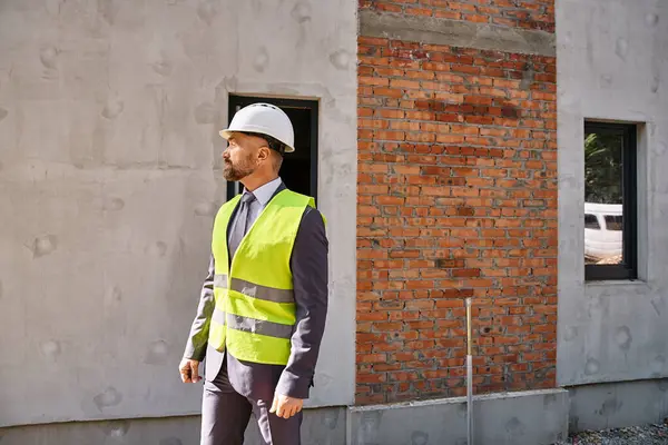 Güvenlik yeleği ve miğferli işadamı inşaat alanında poz veriyor ve başka tarafa bakıyor.