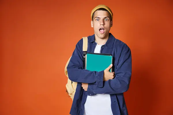 stock image surprised young student with backpack in yellow hat hugging notes on terracotta background