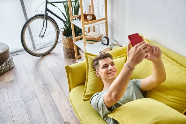 stock image attractive young man with brown hair lying on yellow couch in living room and taking selfie