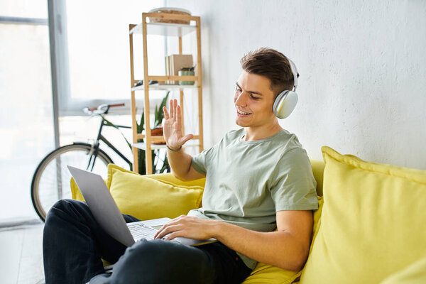 smiling young guy with headphones and laptop in yellow couch saying hello to online meeting sideways