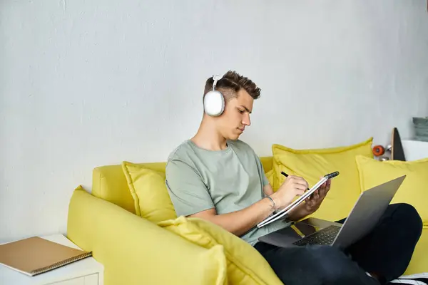 集中注意力的学生 带着耳机和笔记本电脑 躺在黄色沙发上学习写笔记 — 图库照片