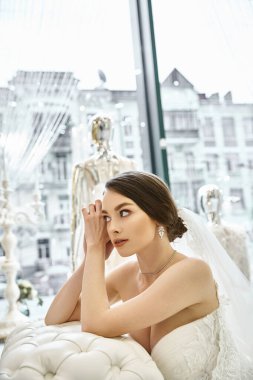A young brunette bride in a beautiful wedding dress sitting peacefully in front of a window in a bridal salon. clipart