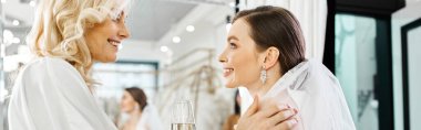 A young brunette bride in a wedding dress stands next to her middle-aged mother in a bridal salon. clipart