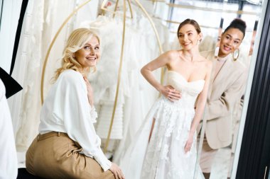 A young brunette bride in a white dress sits on a stool in front of a mirror, admiring her reflection. clipart