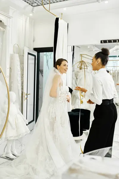一个身穿白衣的年轻新娘和助手站在一起 凝视着他们在新娘沙龙的镜子里的倒影 — 图库照片