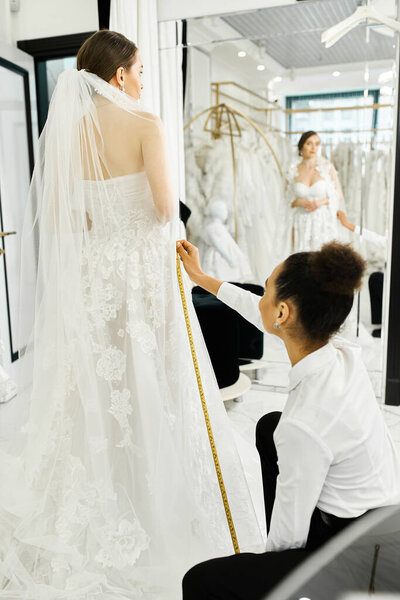 A young brunette bride in a white wedding dress and an African American shopping assistant in a bridal salon.