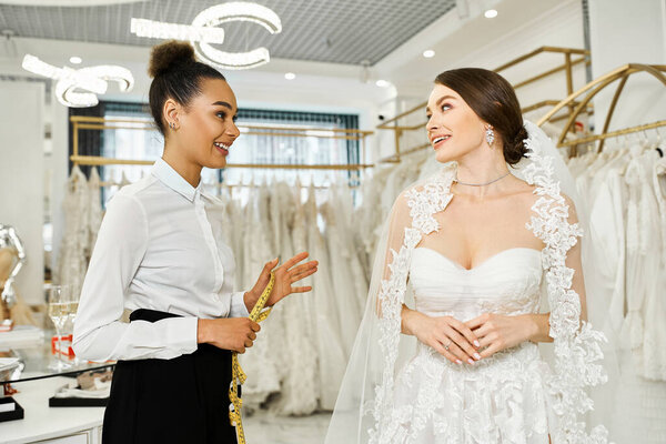A young brunette bride in a wedding dress stands next to an African American shopping assistant in a bridal salon.