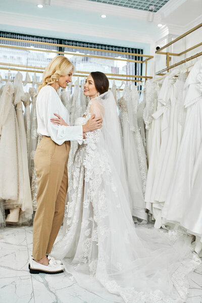 A young brunette bride in a wedding dress and her middle-aged mother stand side by side in a bridal salon, looking at a rack of gowns.