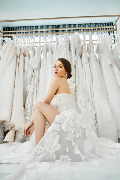 若いブルネットの花嫁がベッドの上に座り ブライダルサロンでウェディングドレスの棚を見つめている — ストック写真