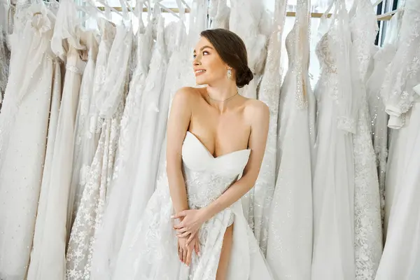A young, beautiful bride stands in front of a rack of white wedding dresses in a bridal salon, carefully selecting her gown.