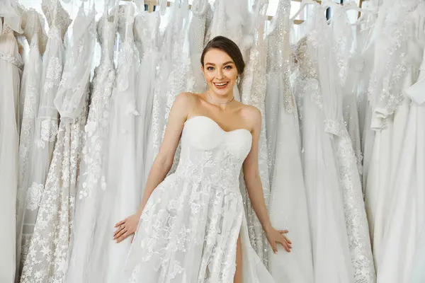 A young brunette bride stands surrounded by a rack of dresses in a wedding salon, looking for her perfect gown.