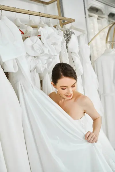 A young, beautiful brunette woman stands in a wedding salon, surrounded by a rack of white dresses.